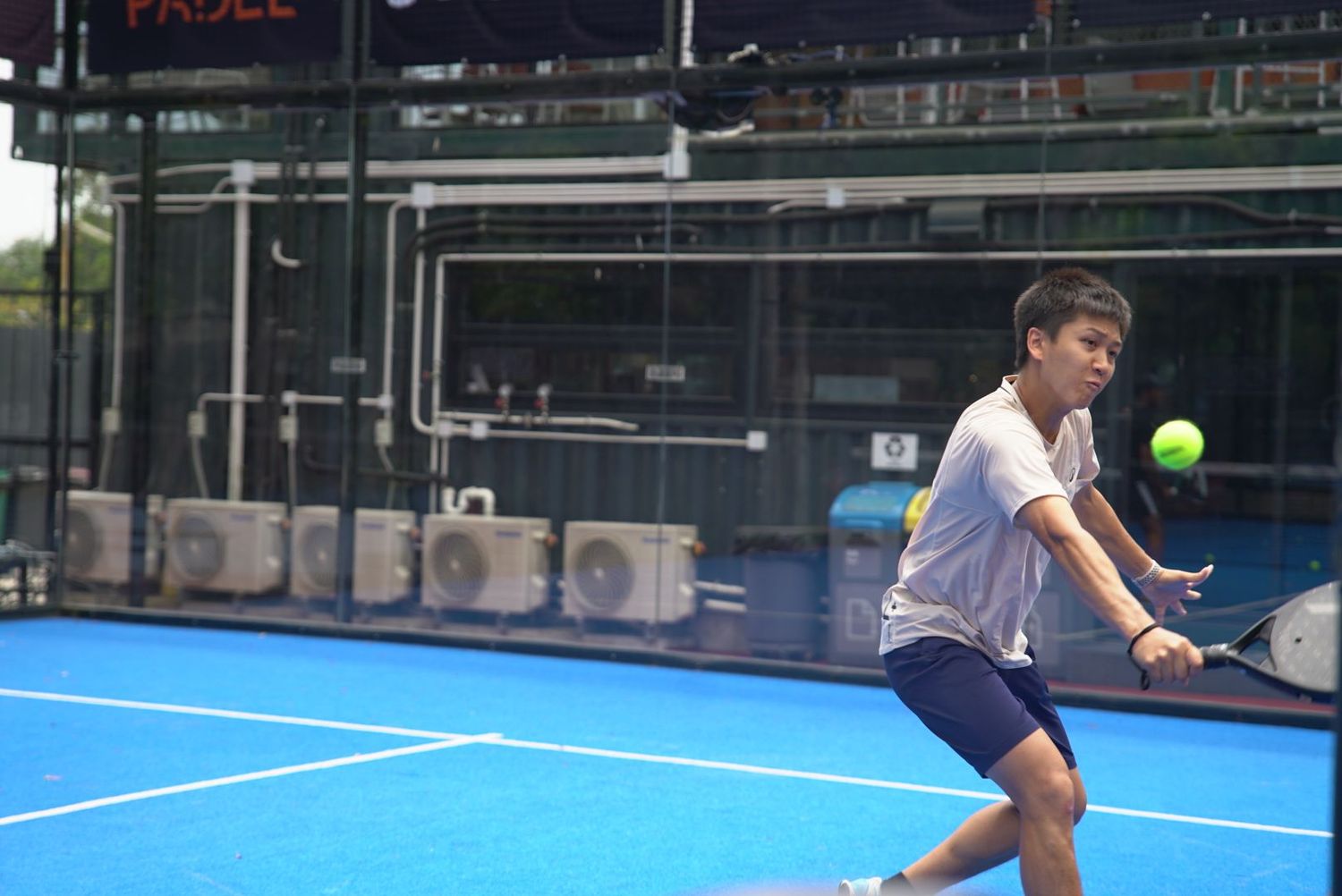 Player hitting a backhand return shot on a bright blue padel court during a match.