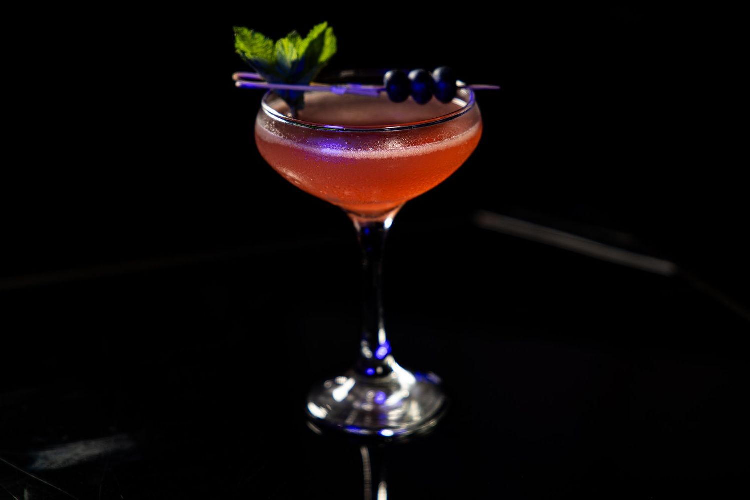 A red cocktail in a coupe glass garnished with purple flowers and herbs against a dark background.