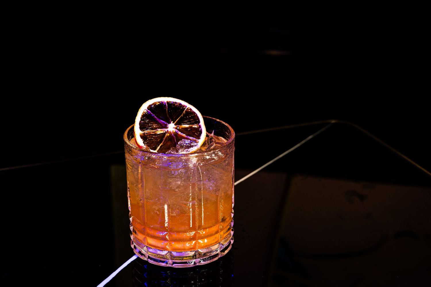 A craft cocktail in a crystal rocks glass garnished with a dehydrated blood orange wheel on a dark background.