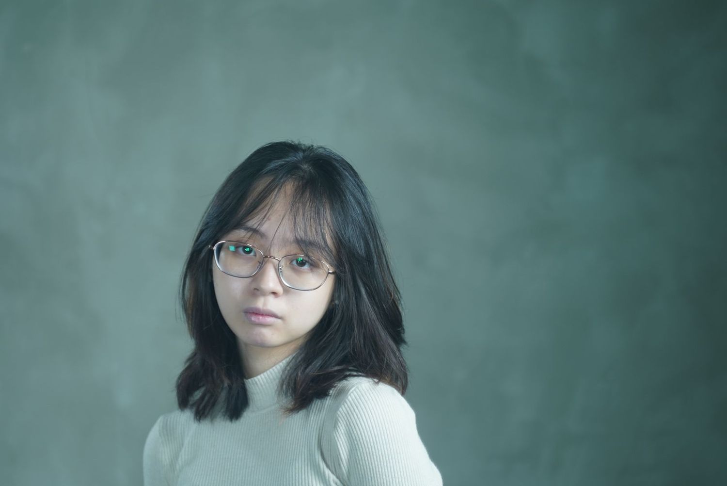 Portrait in soft lighting against green background featuring round glasses and white sweater.