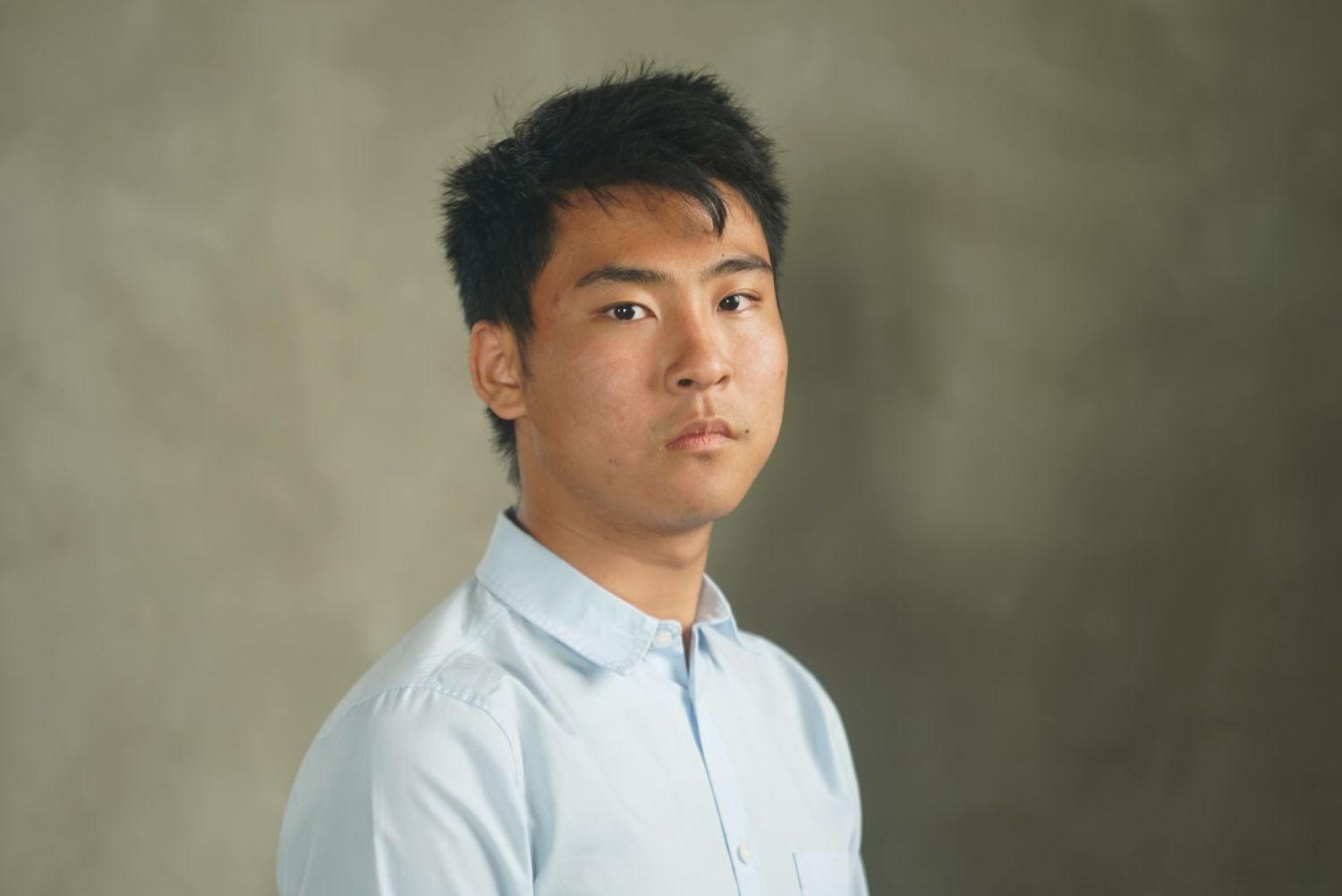 Professional studio portrait with soft lighting features a person in a light blue collared shirt against a muted green background.