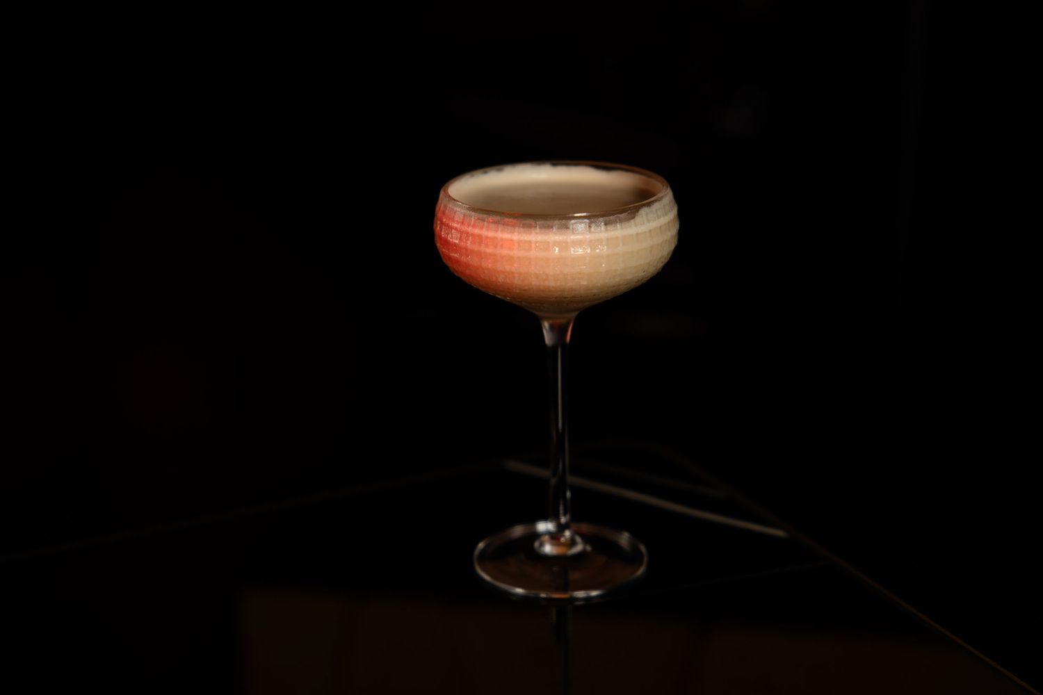 A classic cocktail with pink-orange hue served in a coupe glass against a dark background.