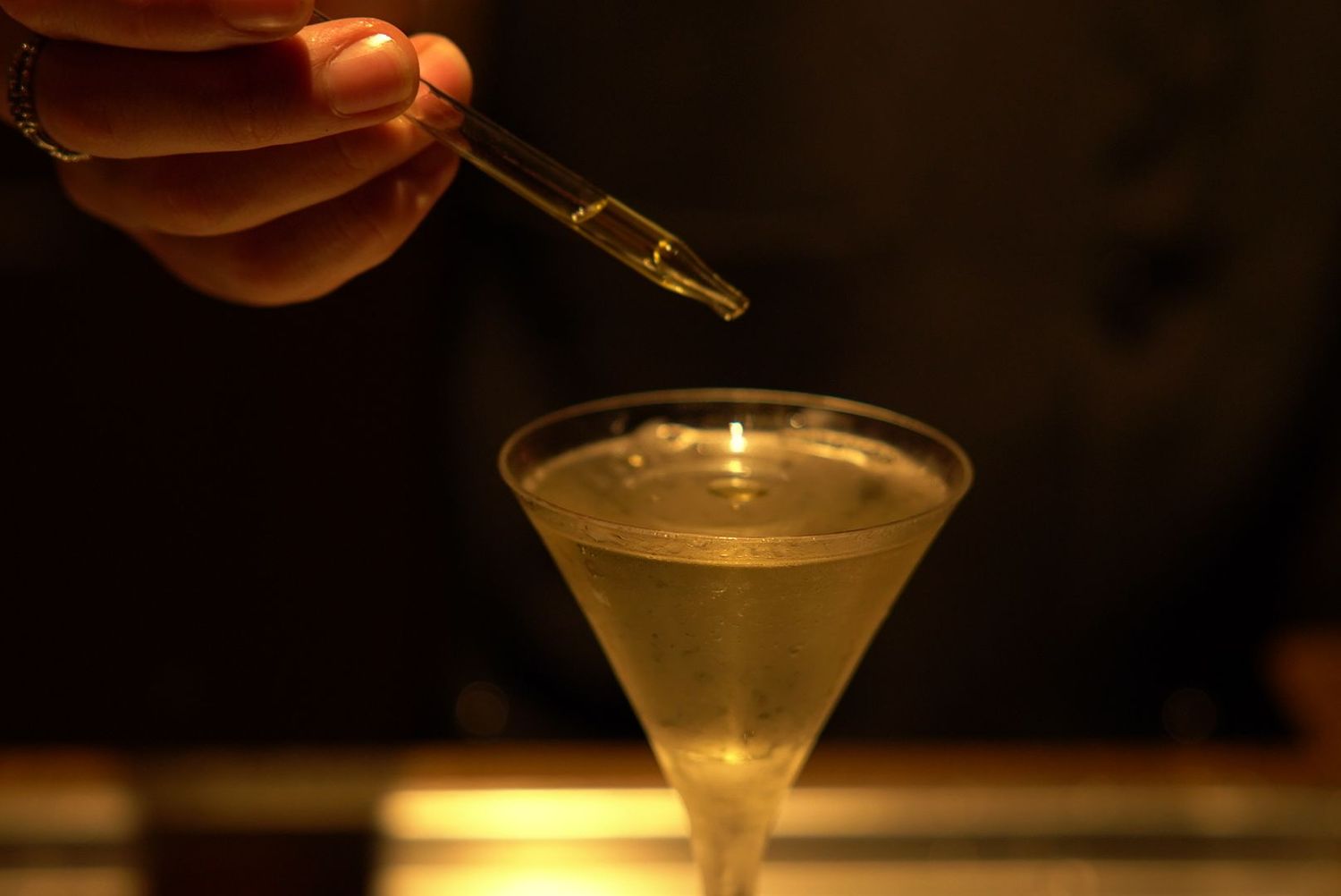 Dropper adding bitters or extract to a chilled martini in atmospheric lighting.