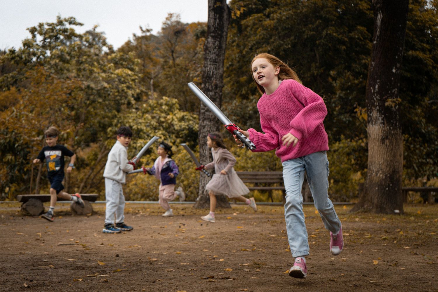 Child in pink sweater playing with toy sword in autumn park setting.
