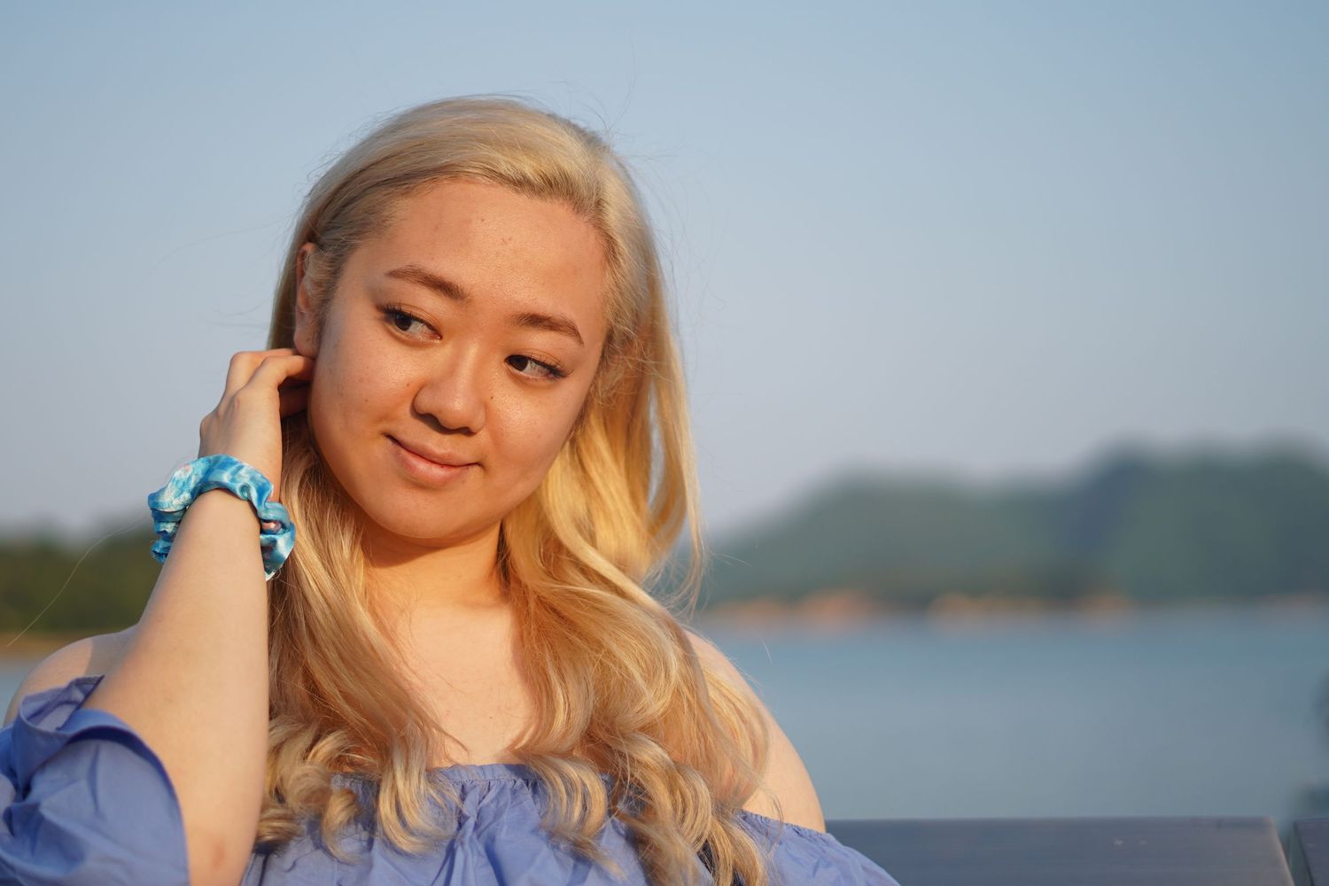 A person with blonde hair and blue top poses outdoors near water during golden hour.