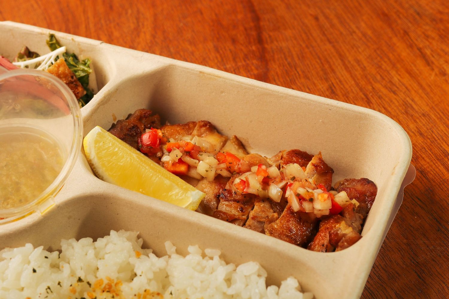 Close-up of a bento box containing white rice, grilled meat with sauce, and a small container of dressing with lemon wedge.