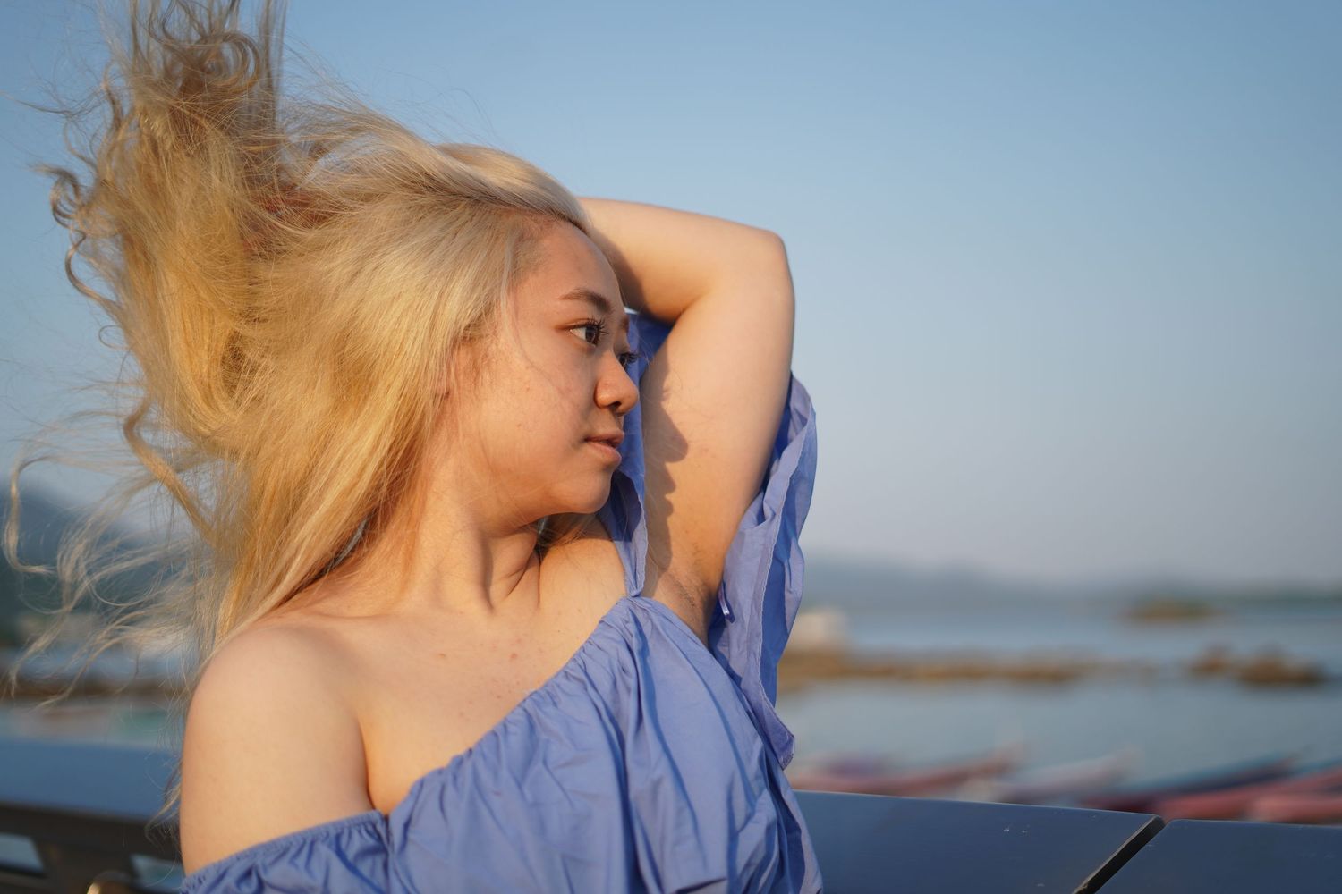 A person with blonde hair in a blue off-shoulder top poses against a serene coastal backdrop at golden hour.