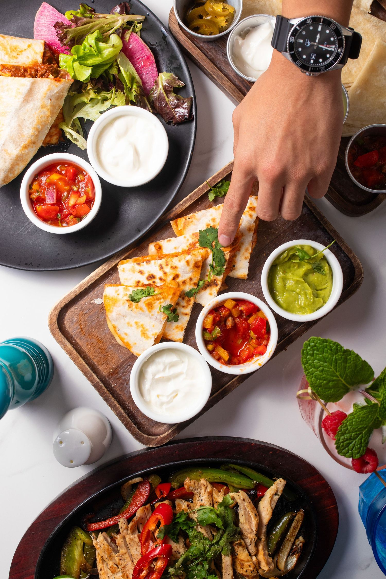 Overhead view of Mexican food spread with quesadillas, dips, salads and colorful drinks on wooden boards and plates.