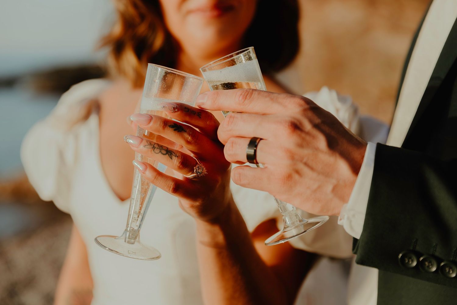 Newlyweds clinking champagne glasses, bride in white dress and groom wearing wedding band, celebrating outdoors.