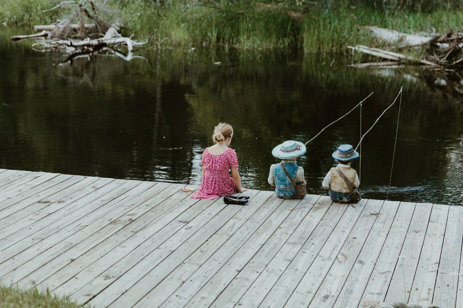 Three children sitting on a wooden dock fishing in a calm pond surrounded by lush green trees.