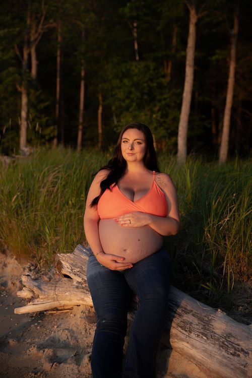 Sunset maternity portrait in coral top against wooded beach background.