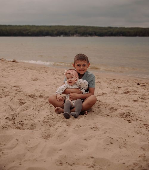 Two figures sitting close together on a sandy beach near a peaceful lake.