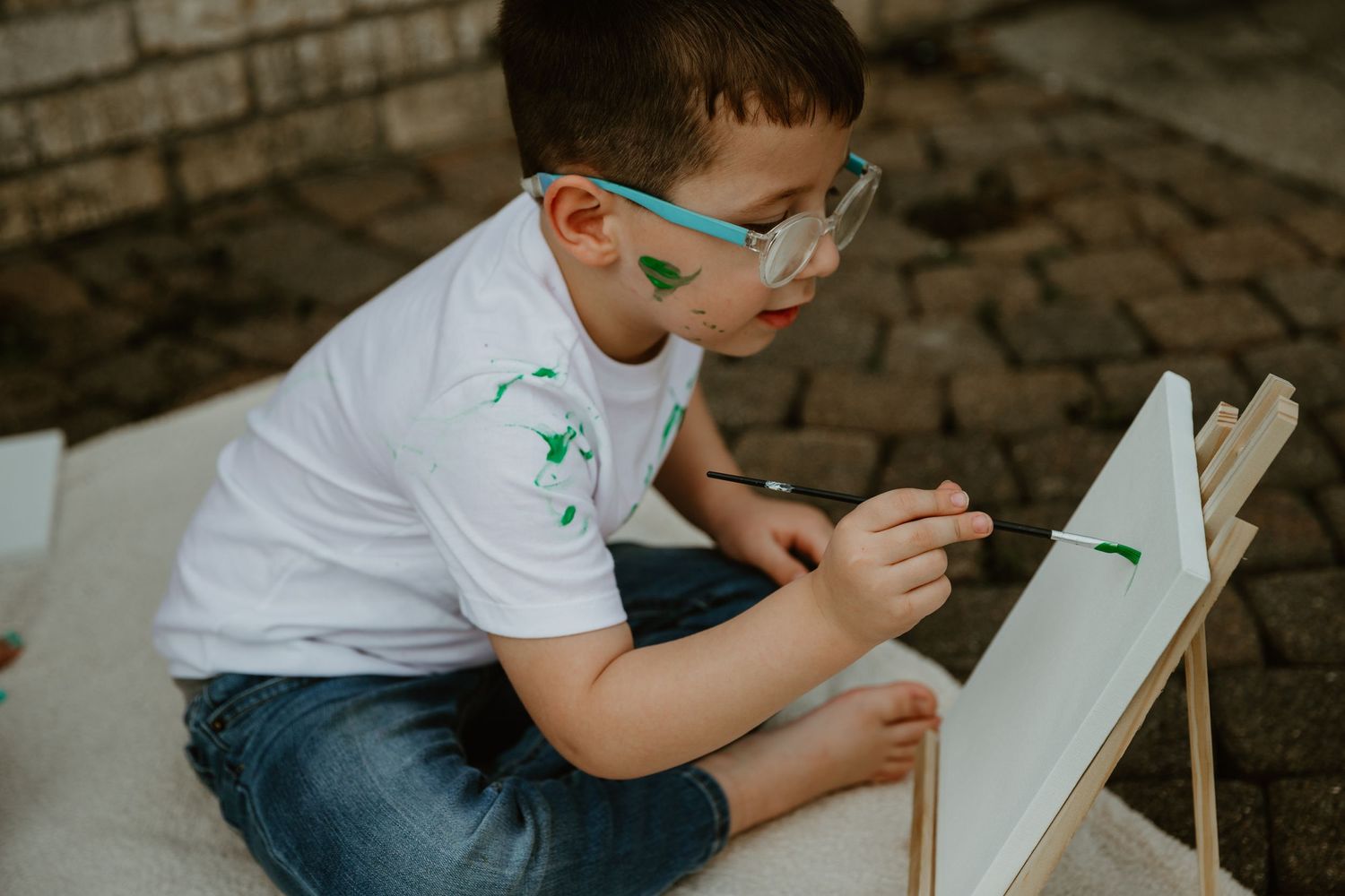 A child in a white t-shirt and glasses sits outdoors while drawing on a white board.
