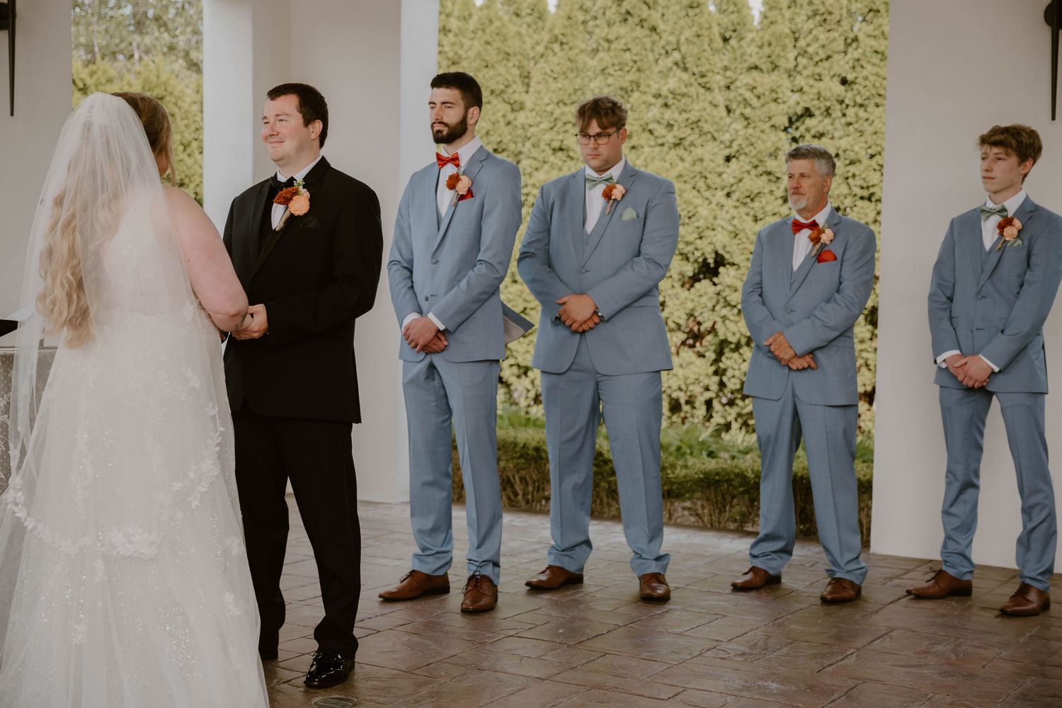 Wedding party lines up under white columns with bridesmaids in red and groomsmen in light gray suits.