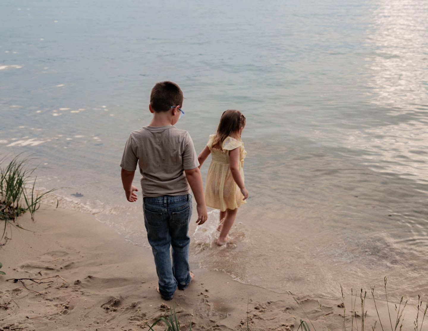 Two children standing at the edge of a sandy beach shore, gazing at calm sunlit water together.