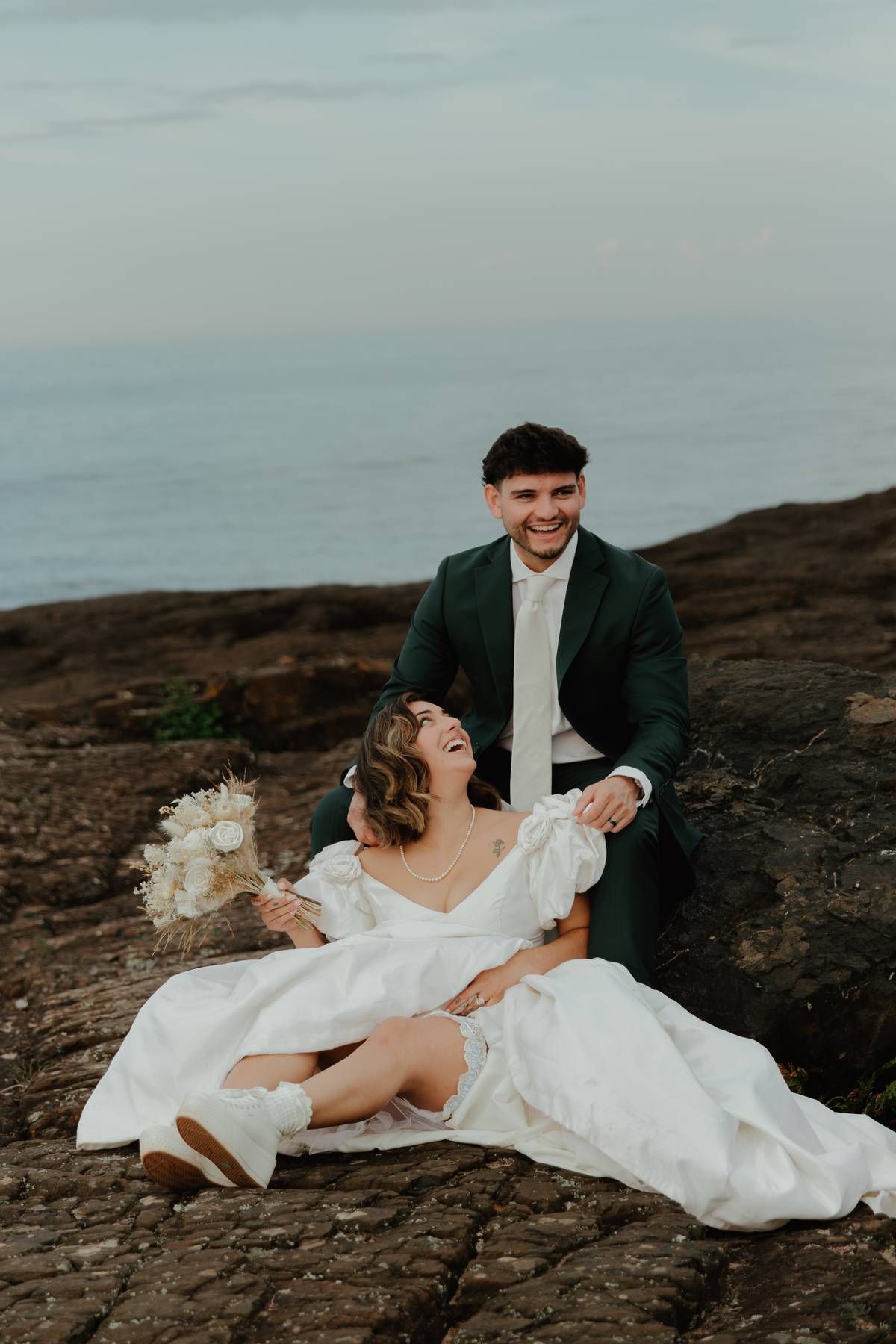 Bride in white gown laughs joyfully as groom in green suit holds her on rocky coastal cliffside.