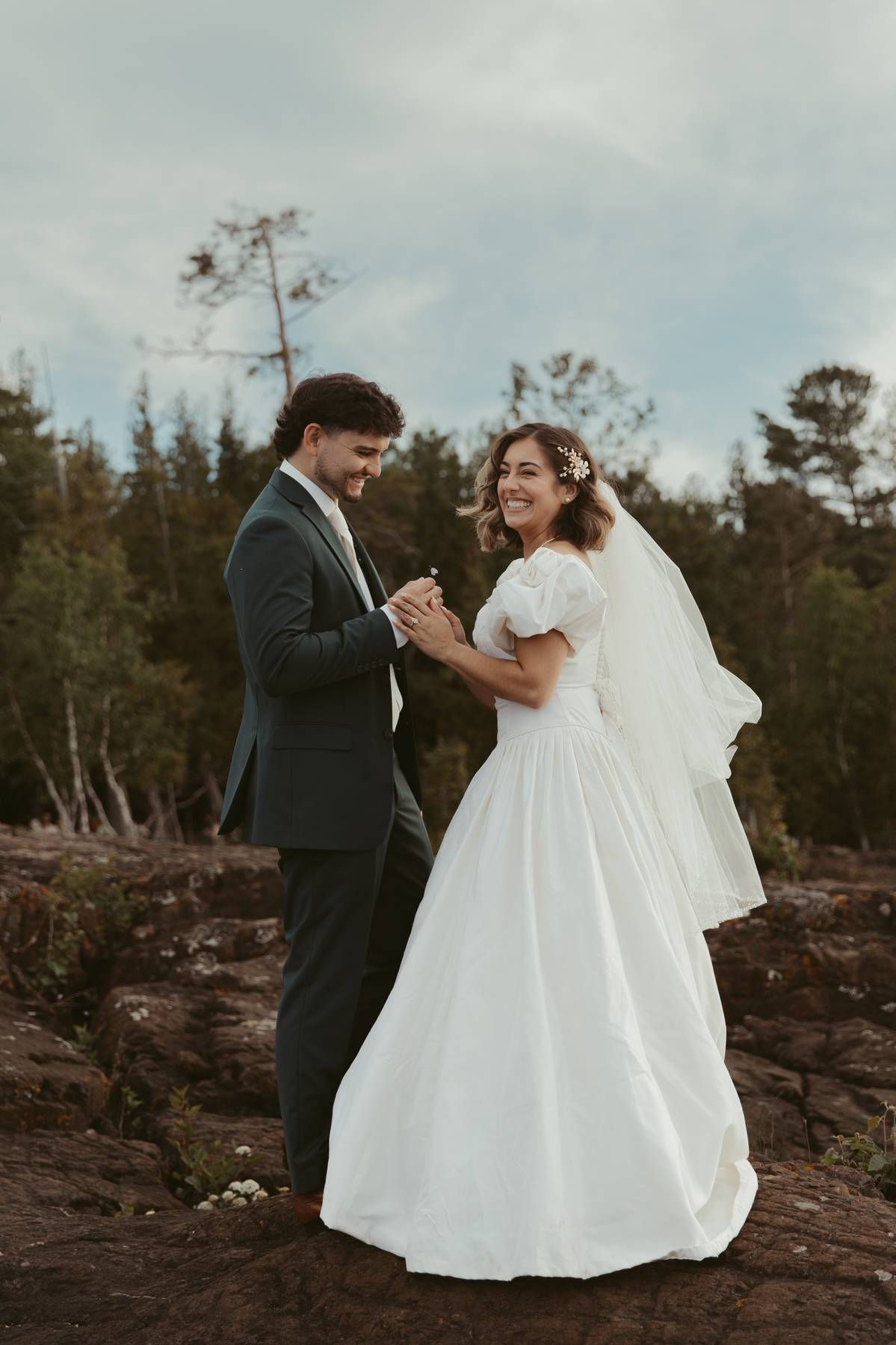 A bride and groom hold hands and smile outdoors on rocky terrain with trees and cloudy sky behind them.