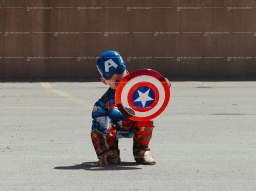 Small child in Captain America costume crouches with iconic shield on concrete ground.