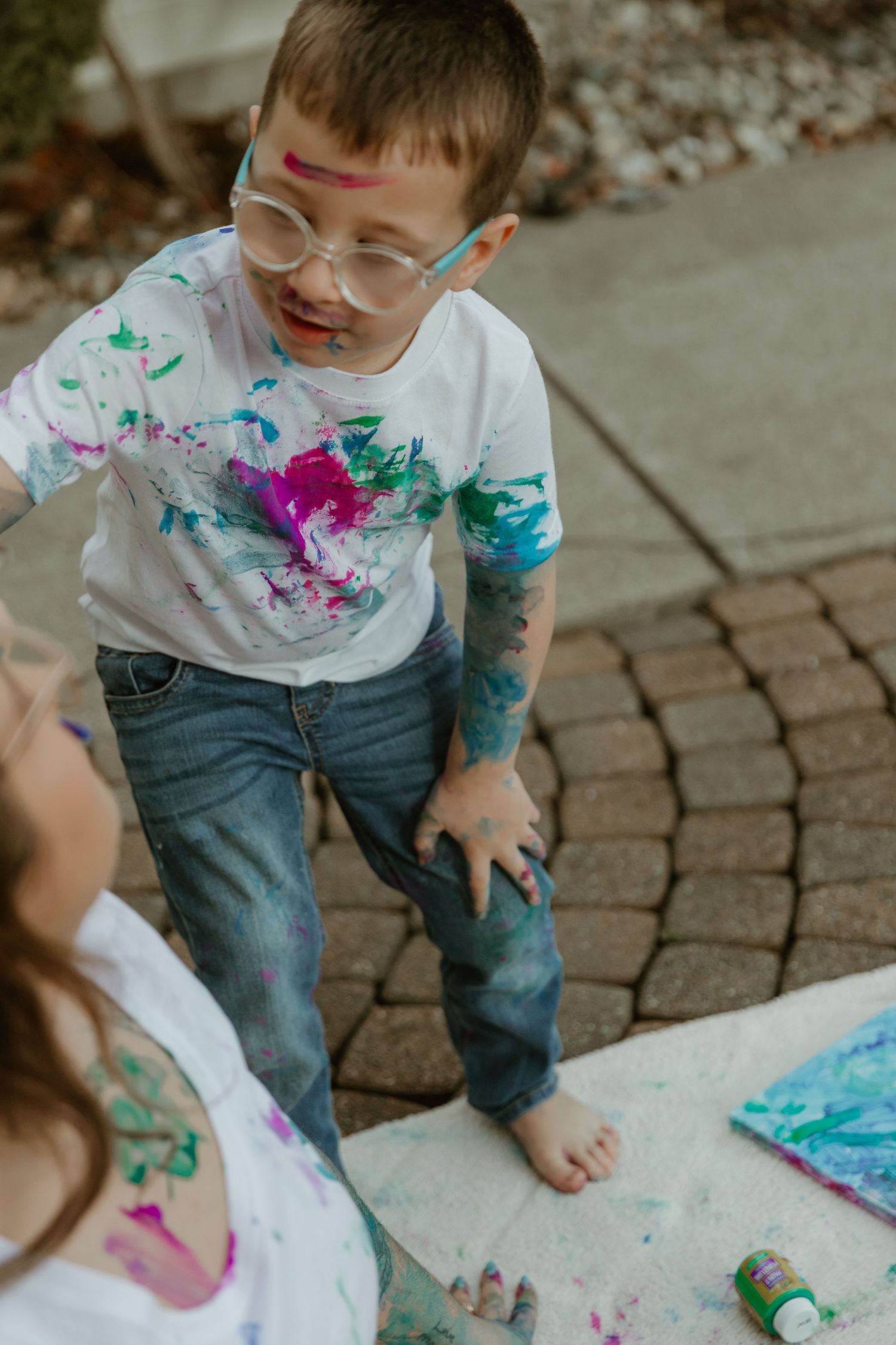 A child in glasses and paint-splattered clothes stands barefoot on a brick patio during a messy art activity.