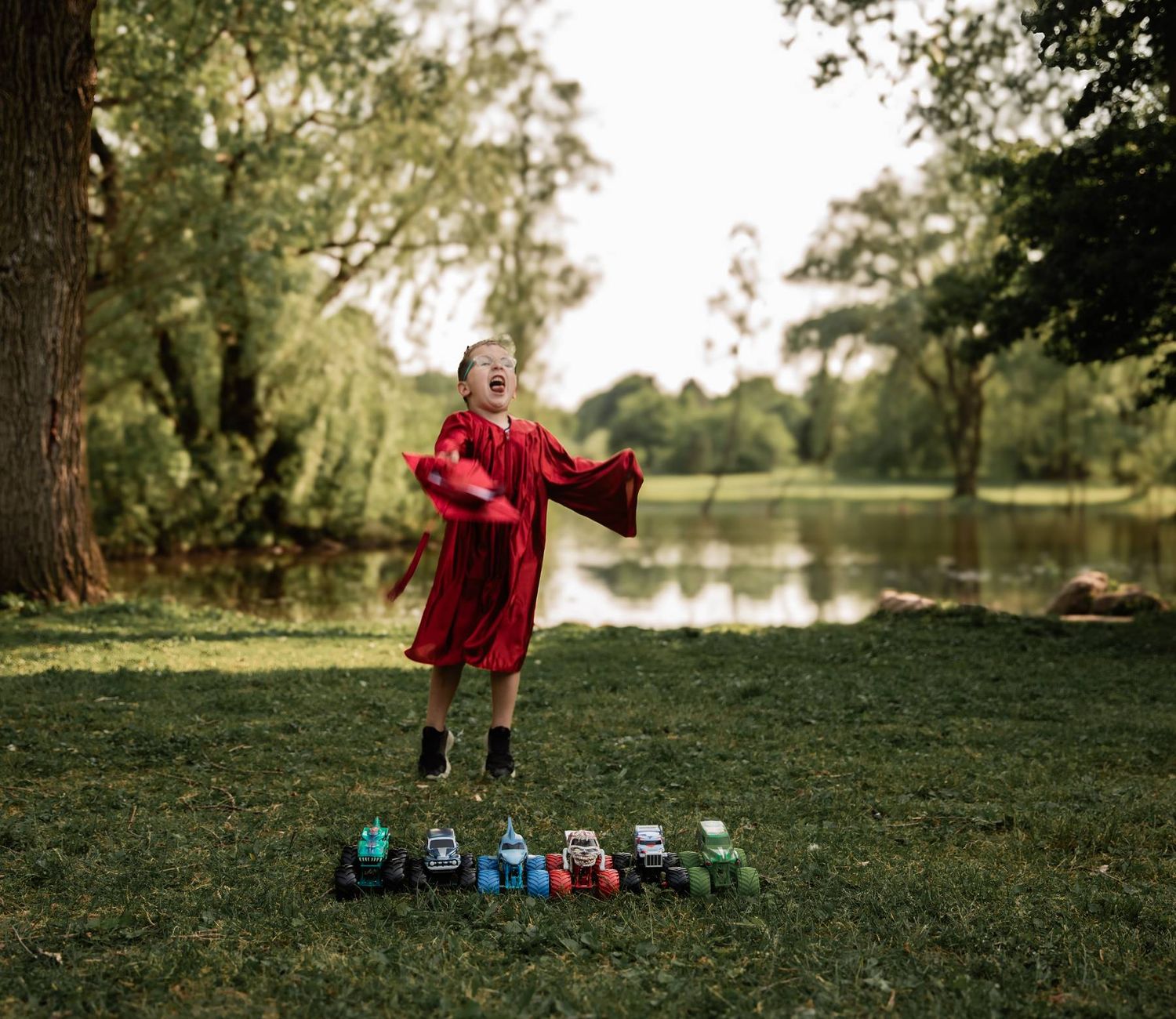 A graduate in red regalia poses playfully with toy tractors lined up on grass by a pond.