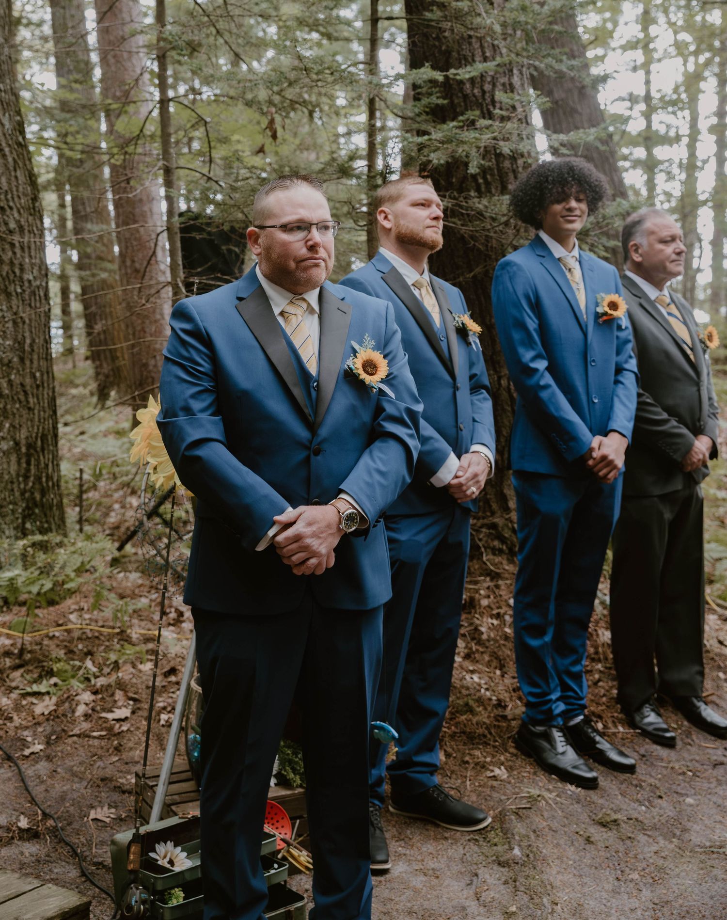 A line of groomsmen in blue suits stand together in a wooded outdoor setting during a wedding ceremony.
