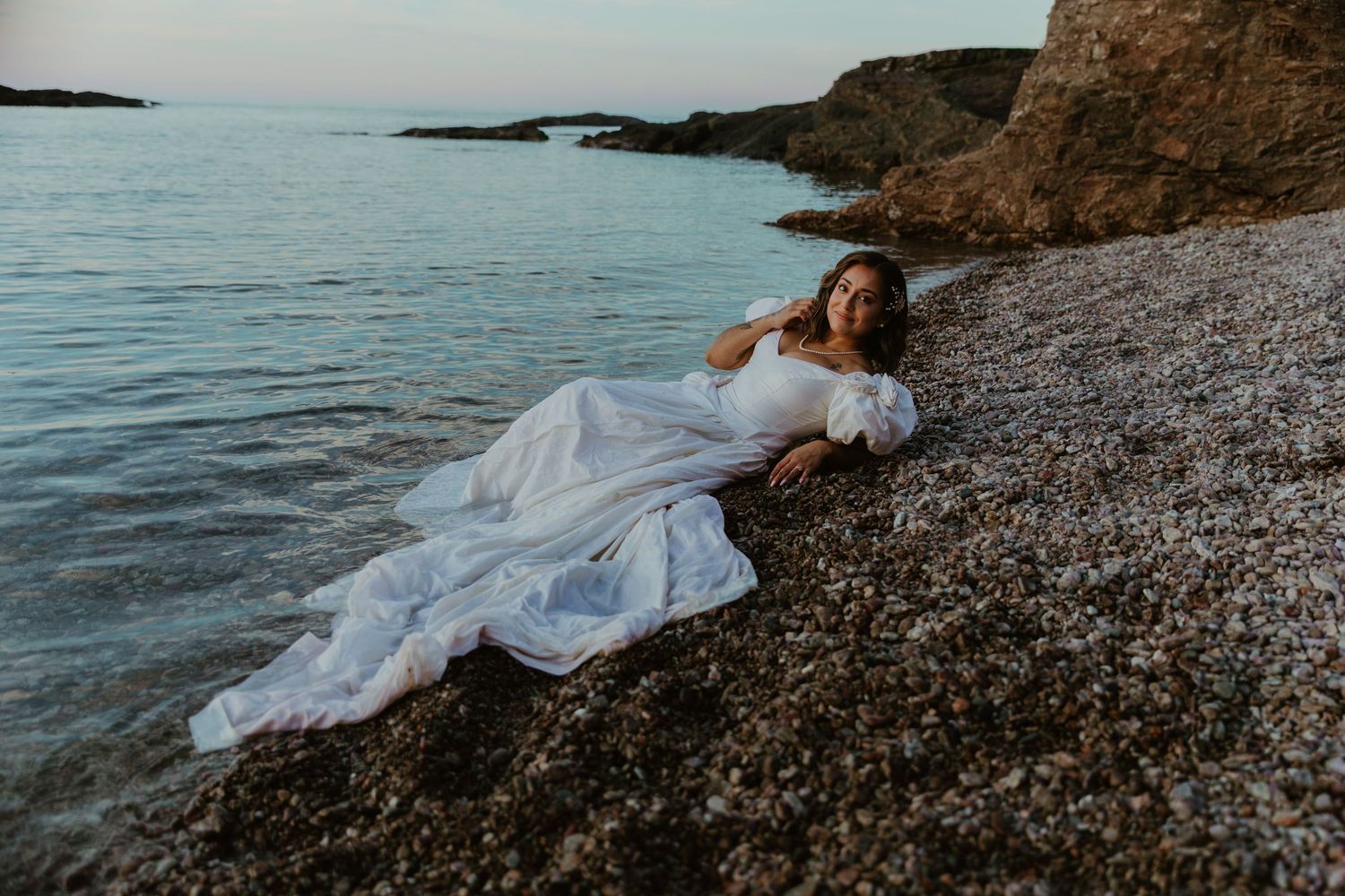 Bride in white wedding gown lying on rocky beach shore at dusk with calm ocean waters in background.
