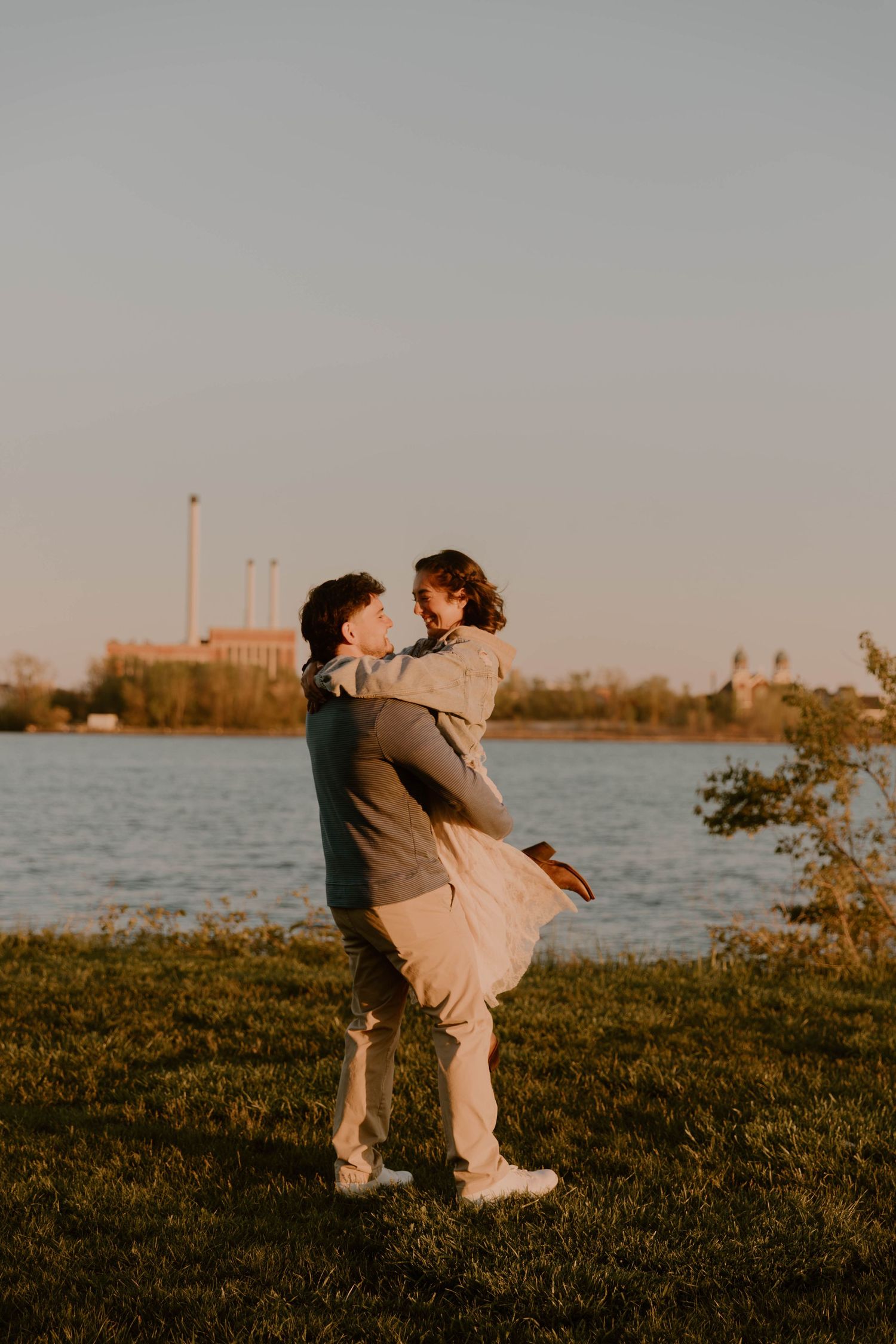 Couple sharing a romantic moment by a lake at golden hour, man lifting woman in a loving embrace outdoors.