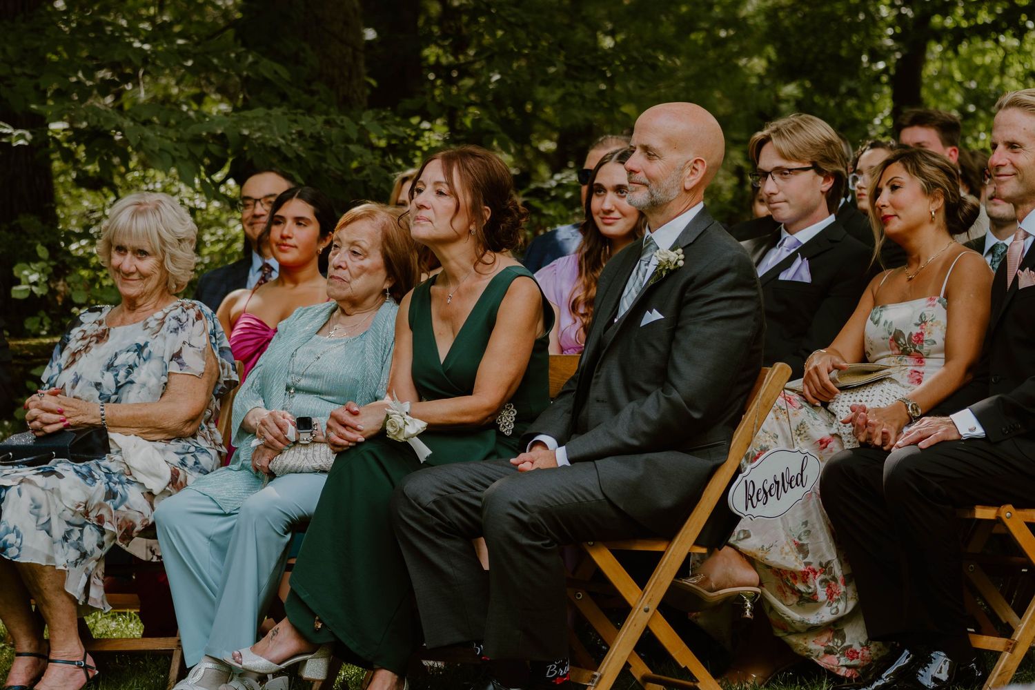 Wedding guests react with joy and laughter while seated in wooden chairs during an outdoor ceremony.
