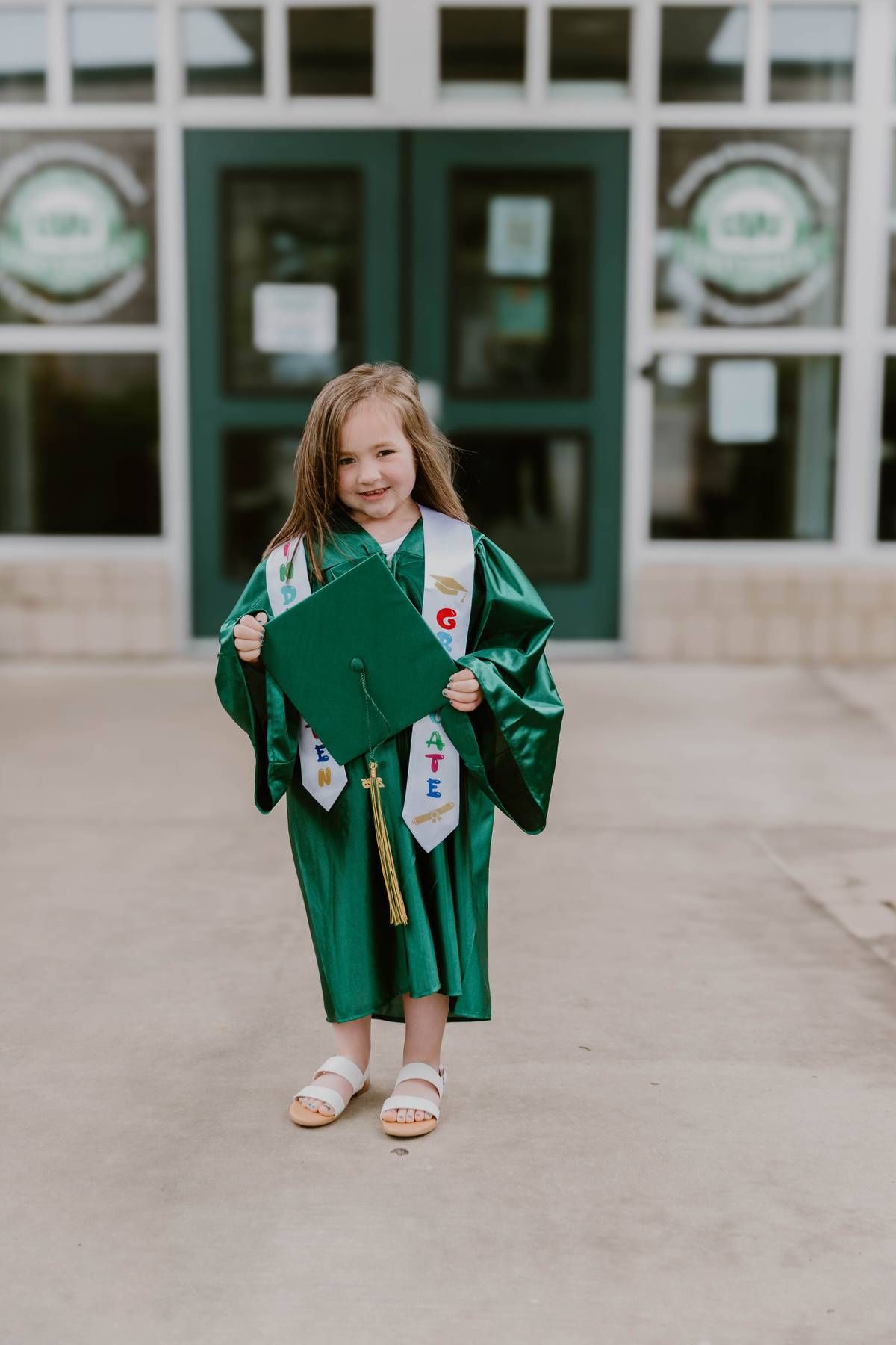 A child in a green graduation gown shows excitement while walking outdoors.