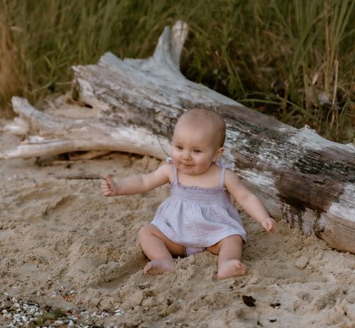 A baby in light colored clothing sitting on a weathered driftwood log.