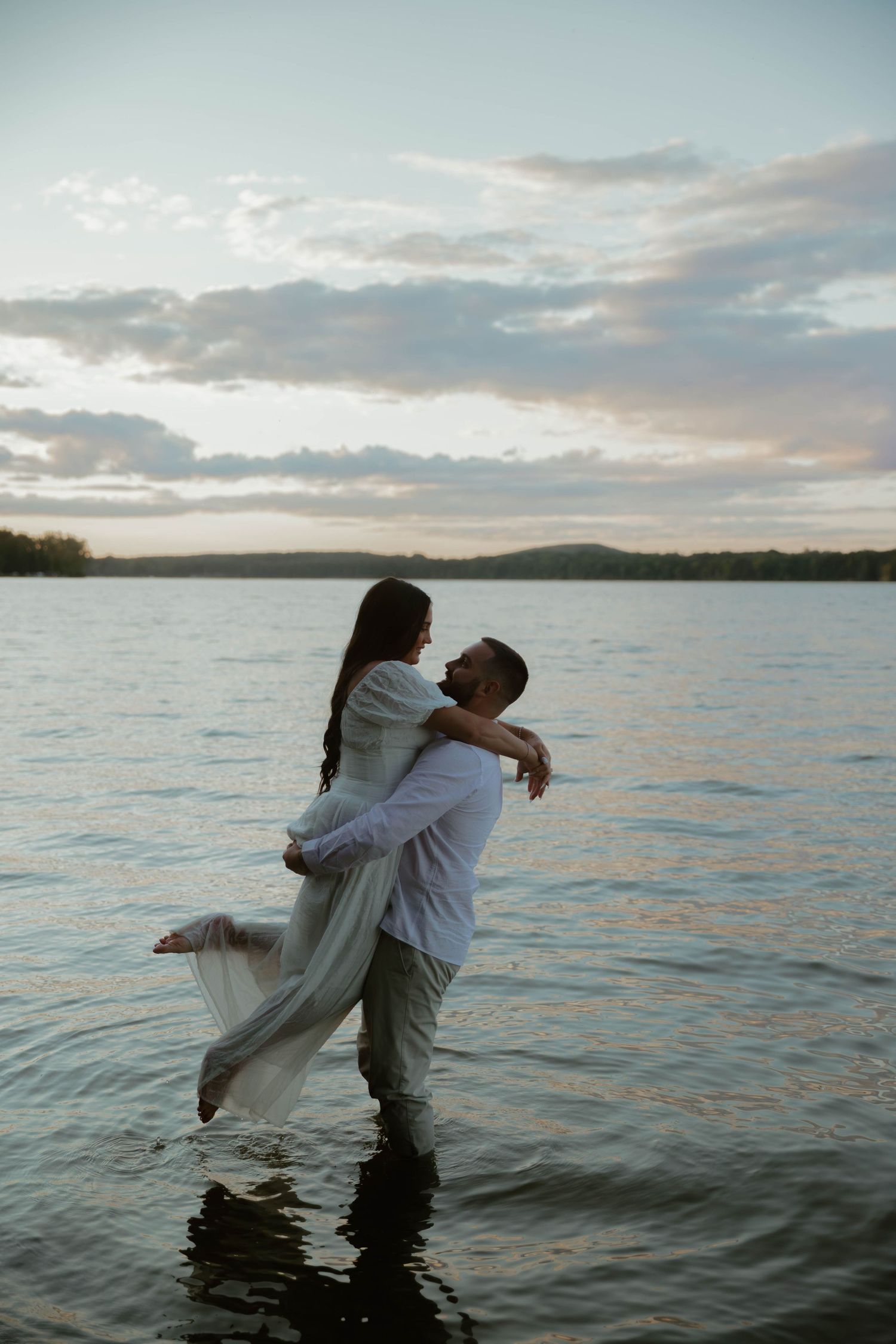 A couple embraces while standing in shallow ocean water at sunset with cloudy skies in the background.