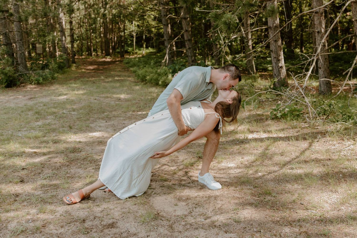 Couple sharing a romantic dip kiss in a sunlit forest clearing, man in blue shirt, woman in white dress.