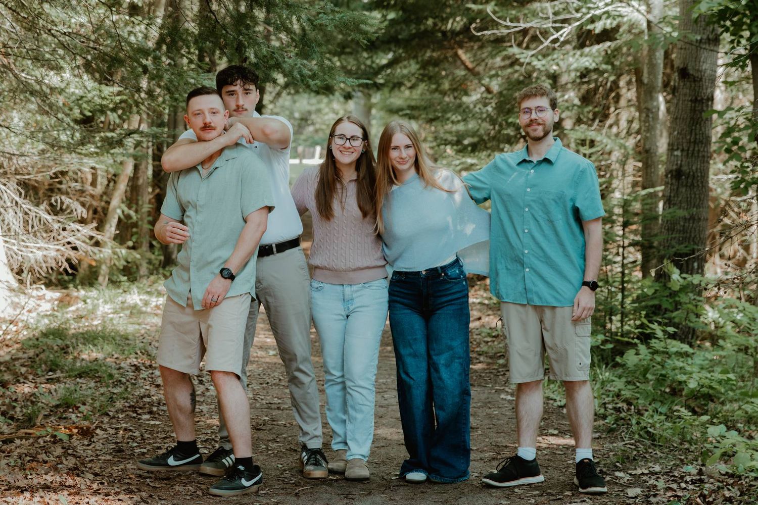 Five people smiling and posing together on a forest trail surrounded by tall green trees.