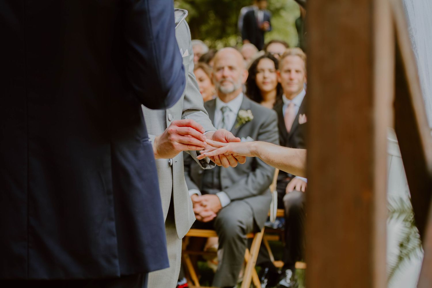 Close-up of hands exchanging rings during an outdoor wedding ceremony with guests seated in the background.