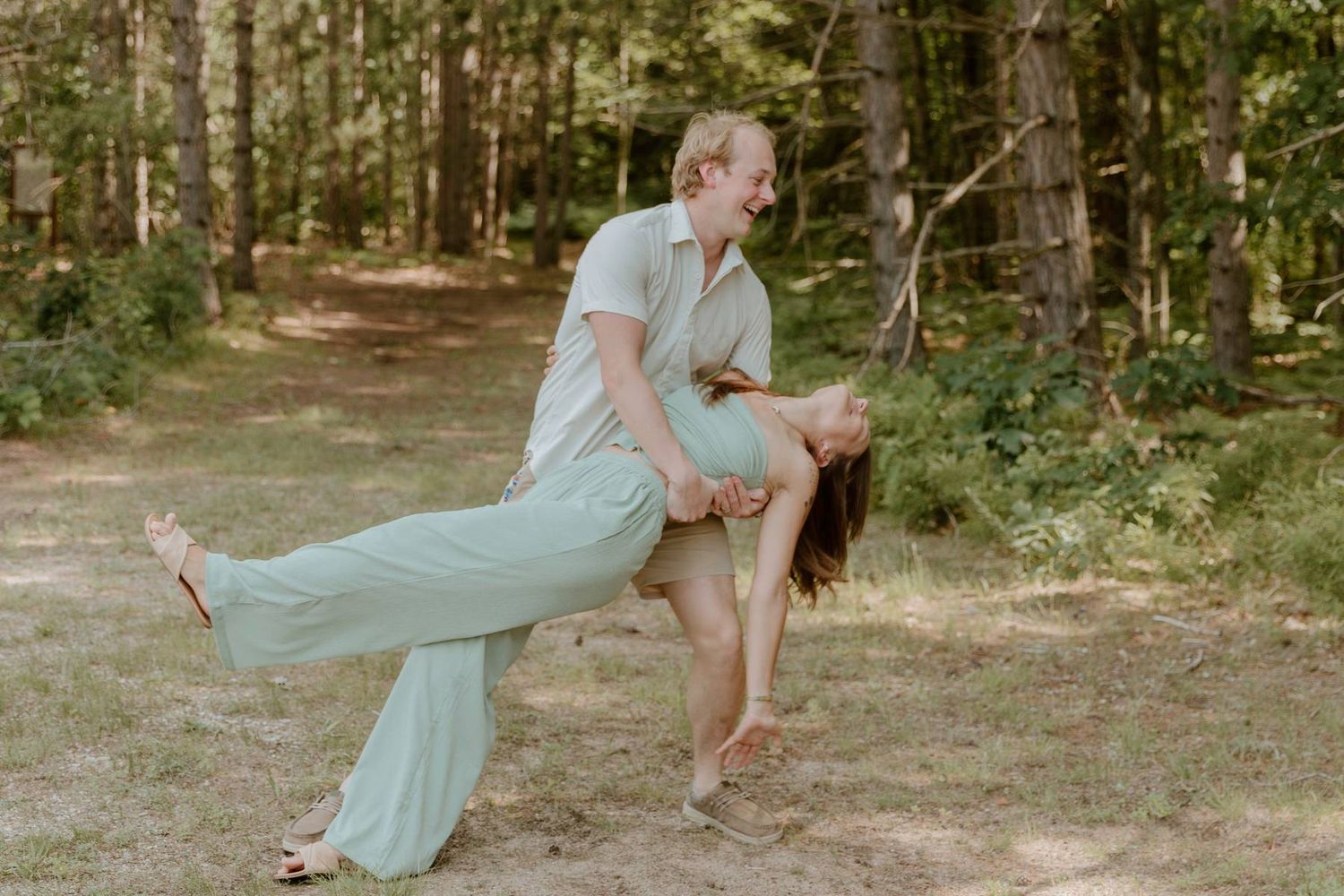 Couple dancing outdoors in forest, man dipping woman in mint green outfit on sandy woodland path.