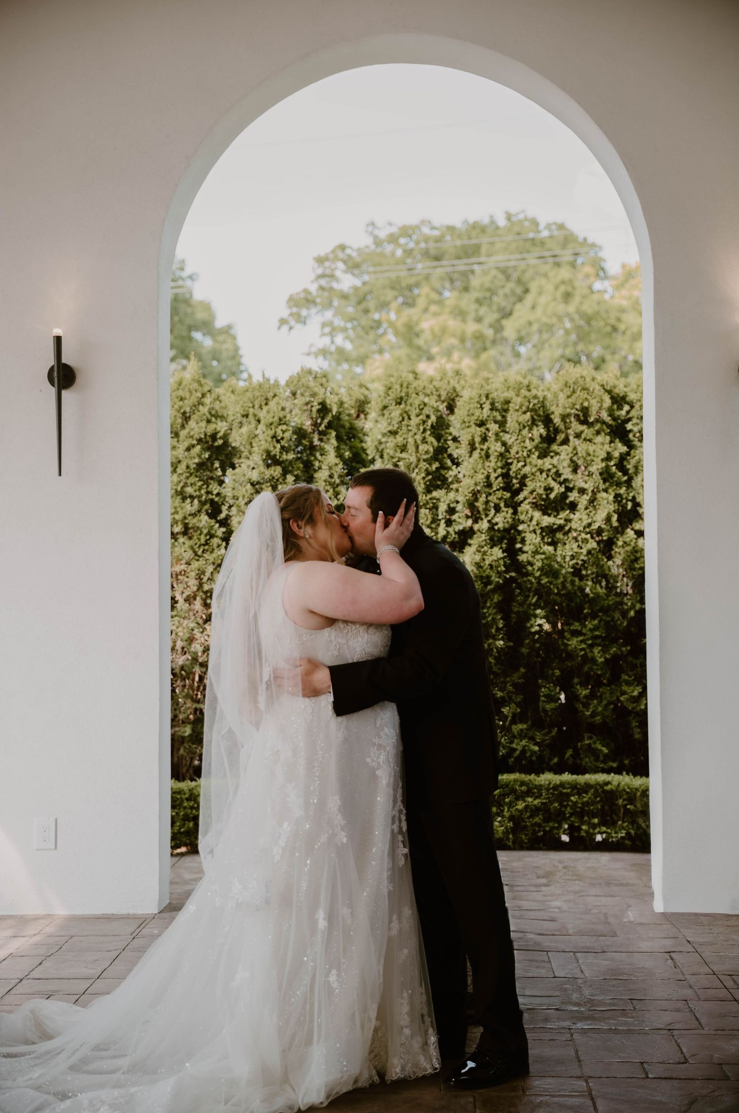 A romantic wedding couple shares a kiss under white stone arches in a garden setting.