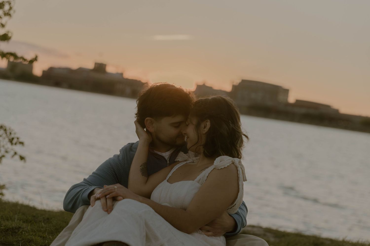 Romantic couple sharing a kiss at sunset by a serene waterfront during golden hour.