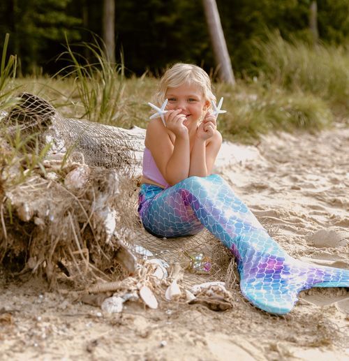 Young person in sparkly mermaid tail costume posing on beach with hands under chin.