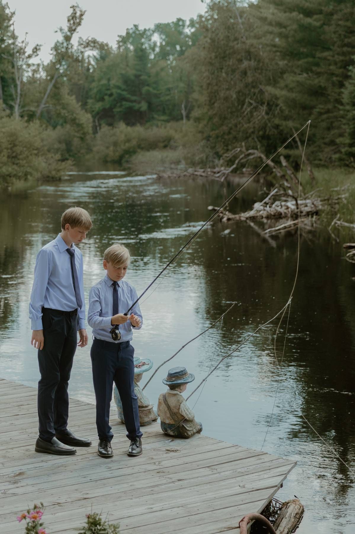 Two boys in dress shirts and ties fish from a wooden dock on a calm river surrounded by lush green trees.