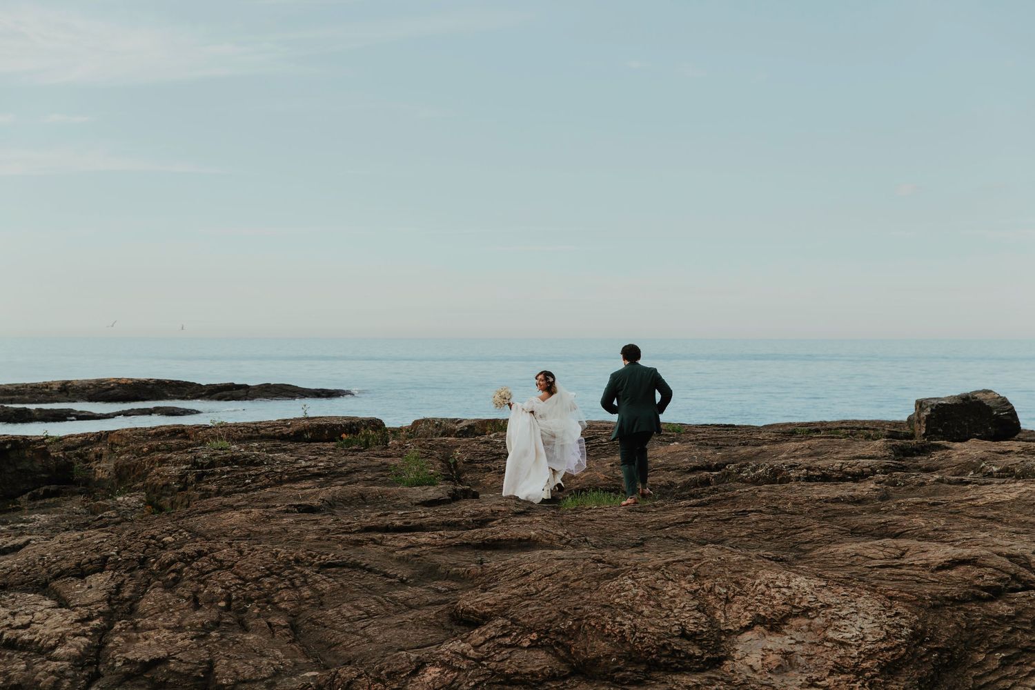A bride and groom walk hand in hand across rocky coastal cliffs toward a serene ocean horizon on their wedding day.