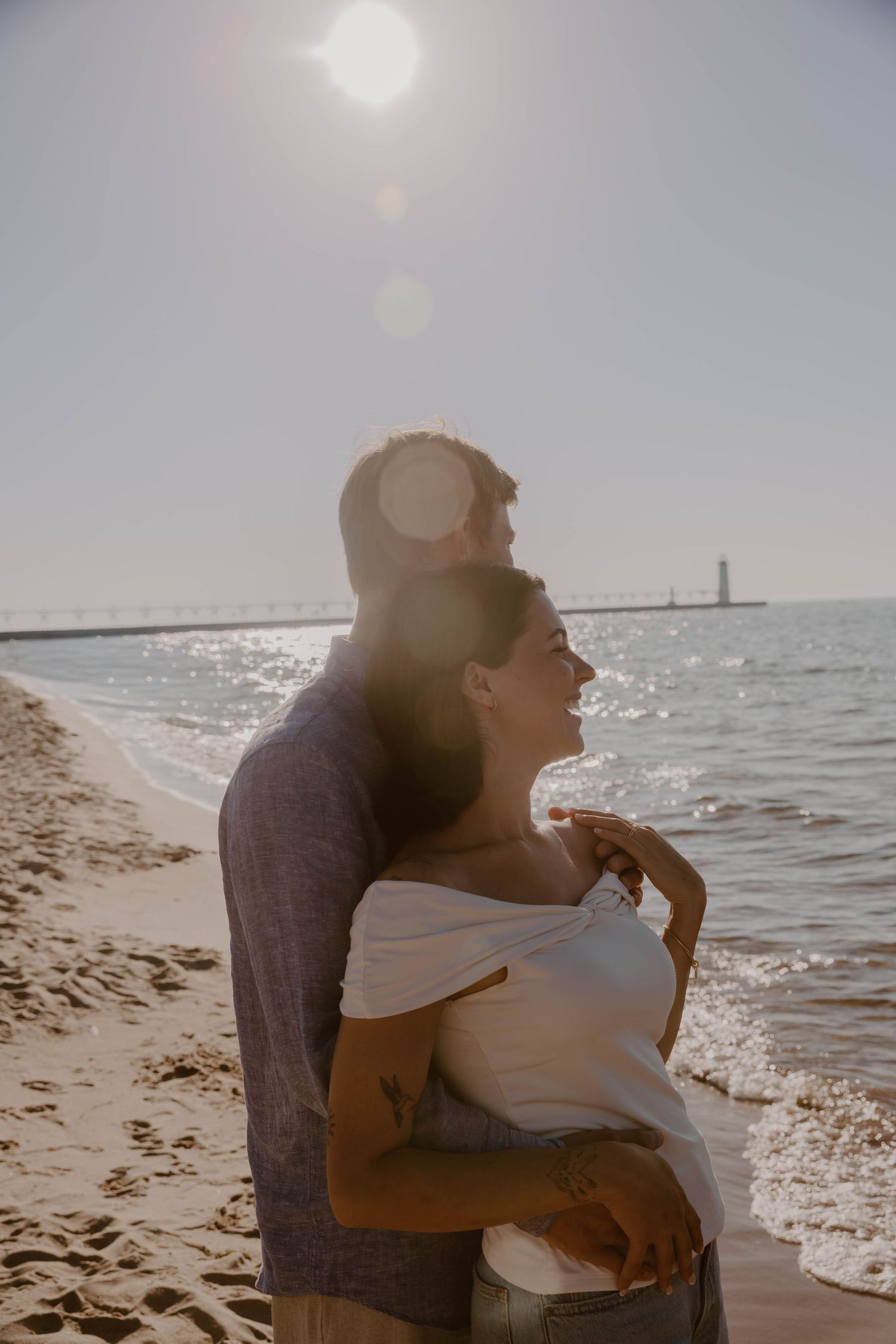 A couple embraces on a sunny beach with a lighthouse visible in the distance across sparkling waters.