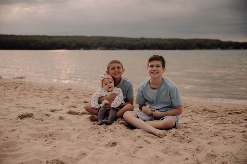 Three figures sit together on a beach with a calm lake and tree-lined shore in the distance.