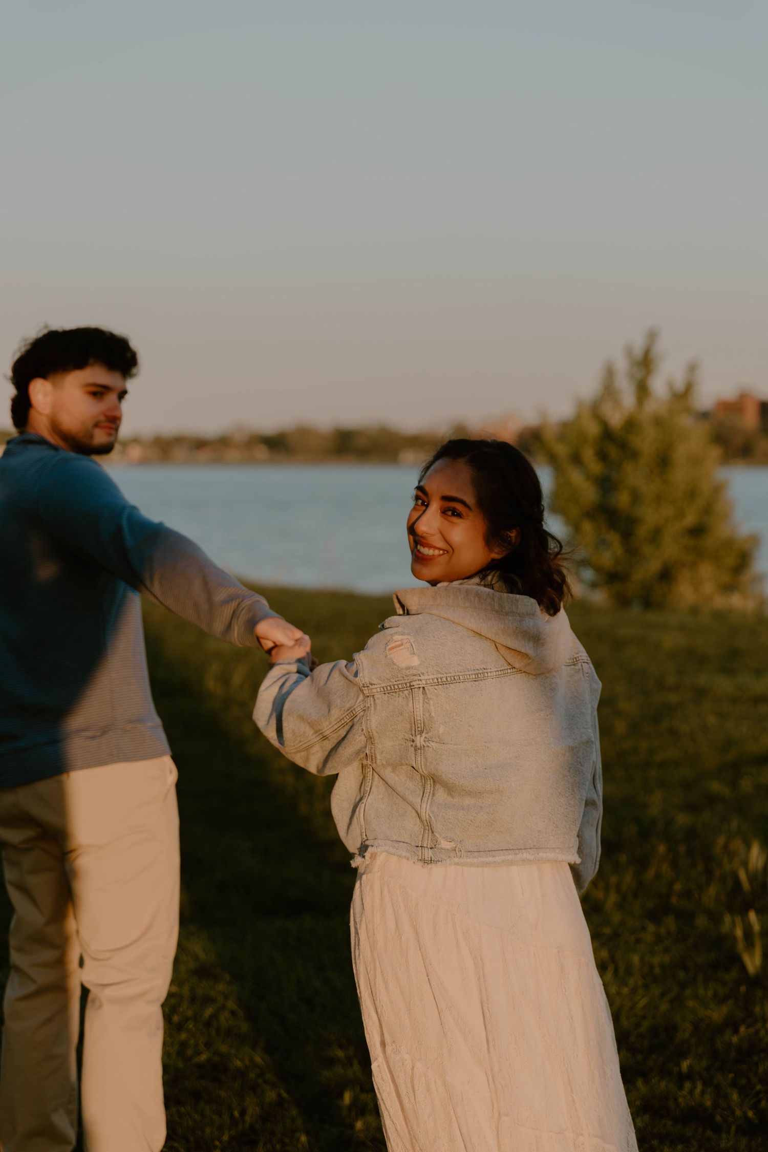 Couple holding hands at sunset near a lake, woman in denim jacket smiling over her shoulder.