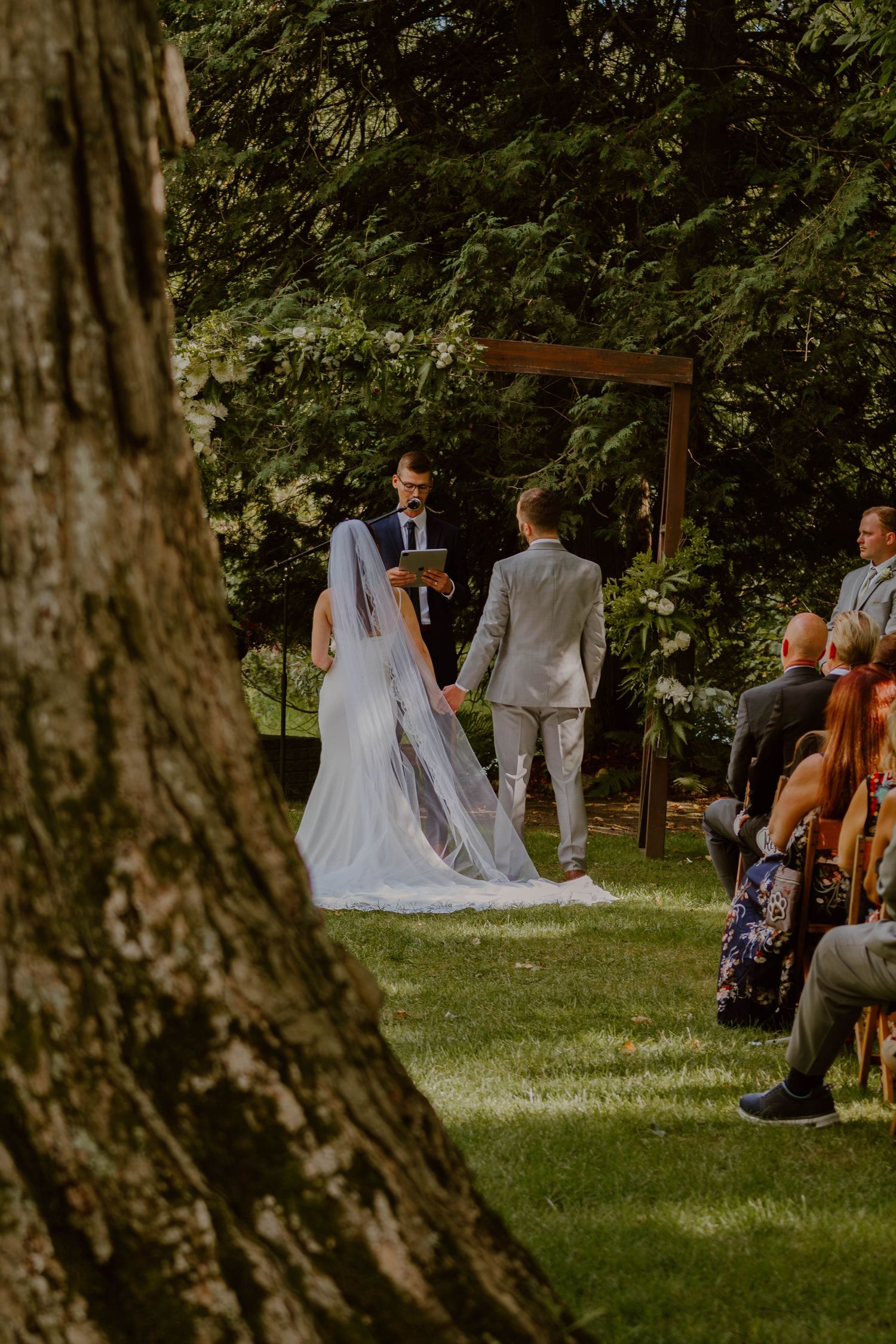 An outdoor wedding ceremony in a wooded setting with guests seated and a wooden arch framing the couple.