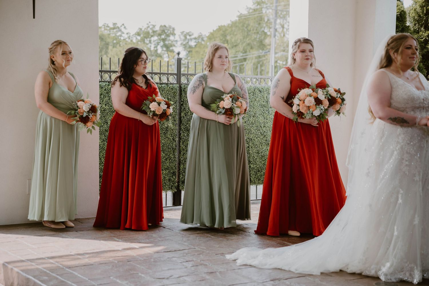 Bridesmaids in alternating sage green and red floor-length dresses stand beside bride in white wedding gown.