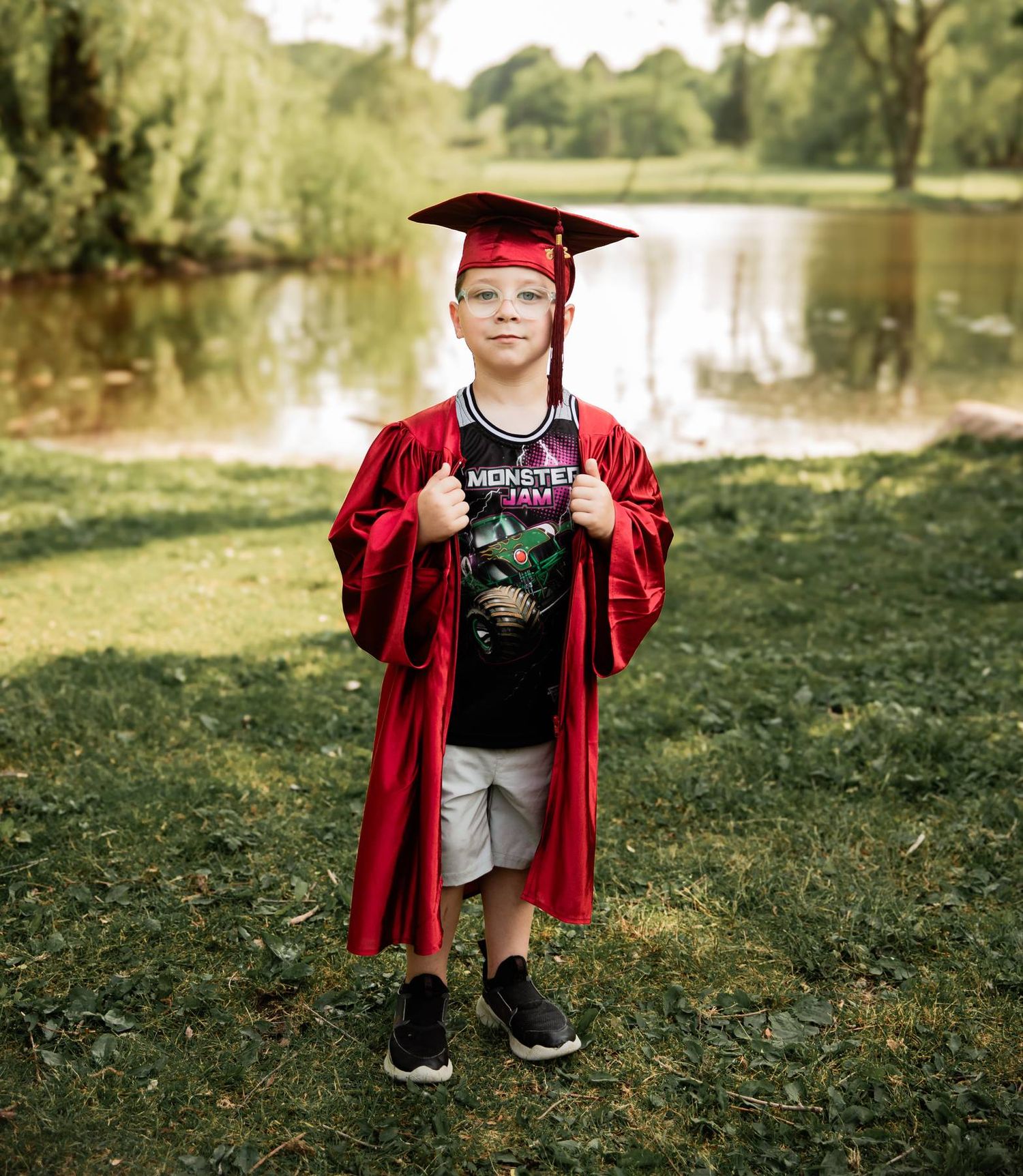 Graduate posing in red graduation gown and cap near a scenic pond in a park setting.