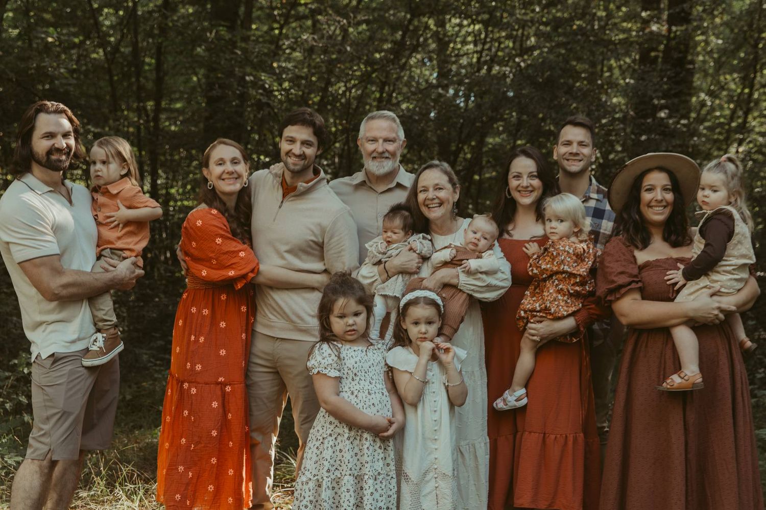Large family group portrait with adults and children smiling outdoors in a lush green forest setting.