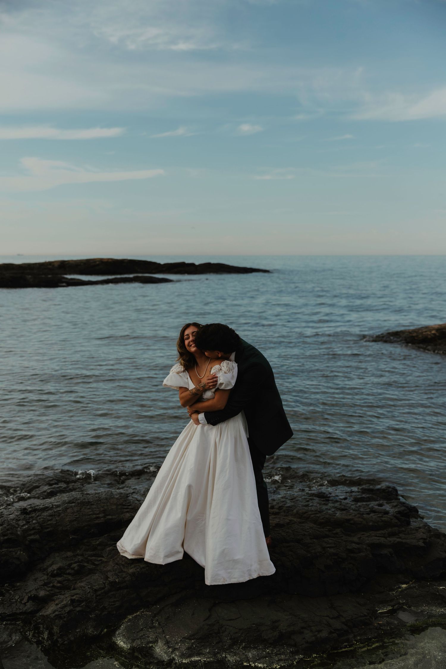 A bride in a white gown embraces her groom on rocky coastal shores with calm blue ocean and sky backdrop.