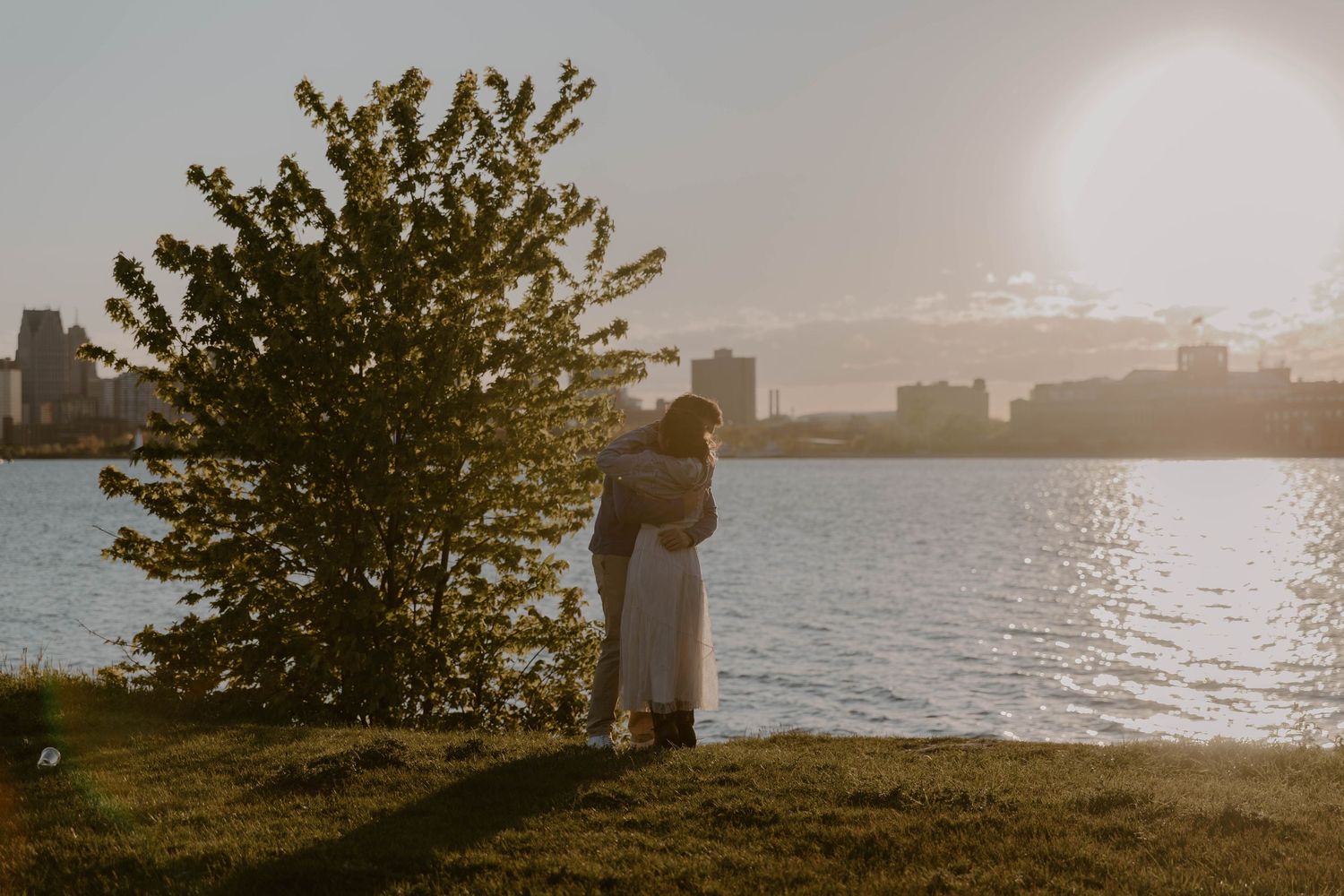 Couple embracing by waterfront at golden hour with city skyline and tree in background.