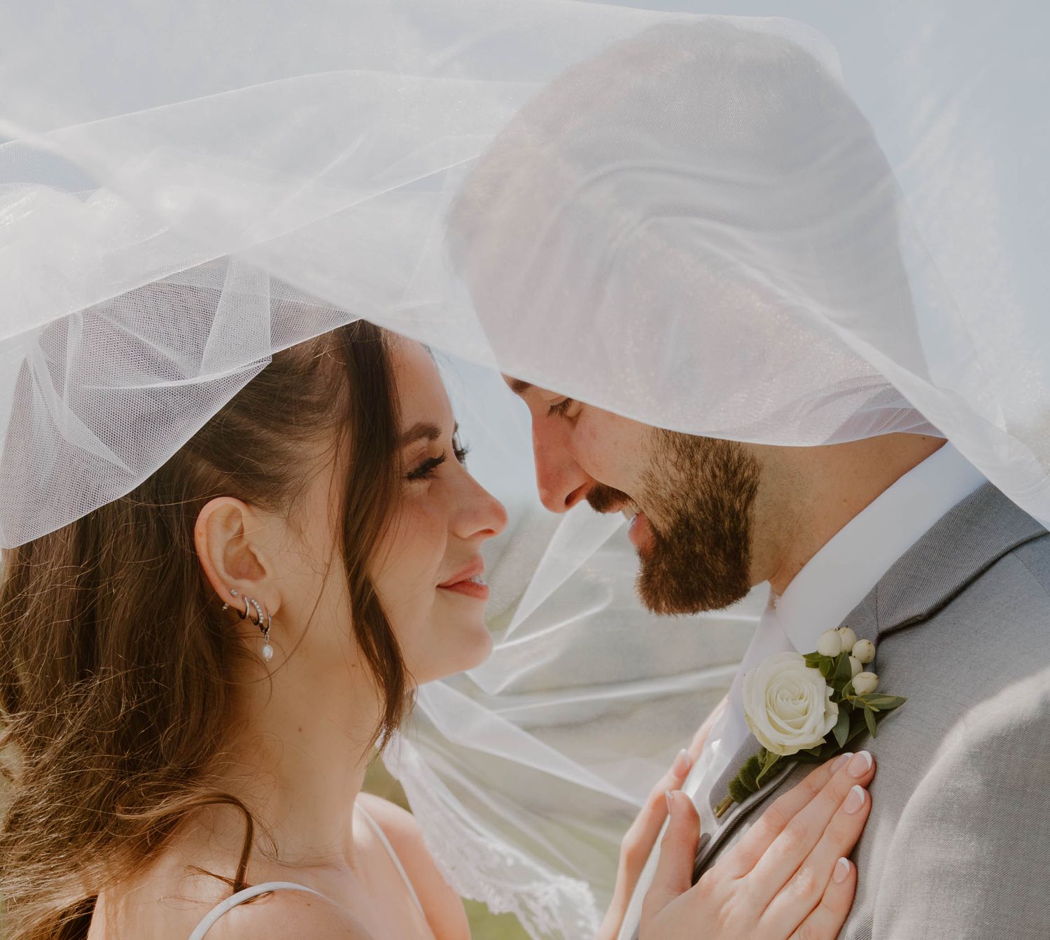 Newlyweds embrace tenderly under a delicate white veil during their outdoor wedding photoshoot.
